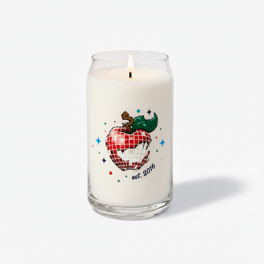 A white wax candle in a clear glass jar with a lit wick, featuring a graphic of a red-and-white checkered apple with a green leaf and the text 'est. 2016' on the front, photographed on a clean white background.