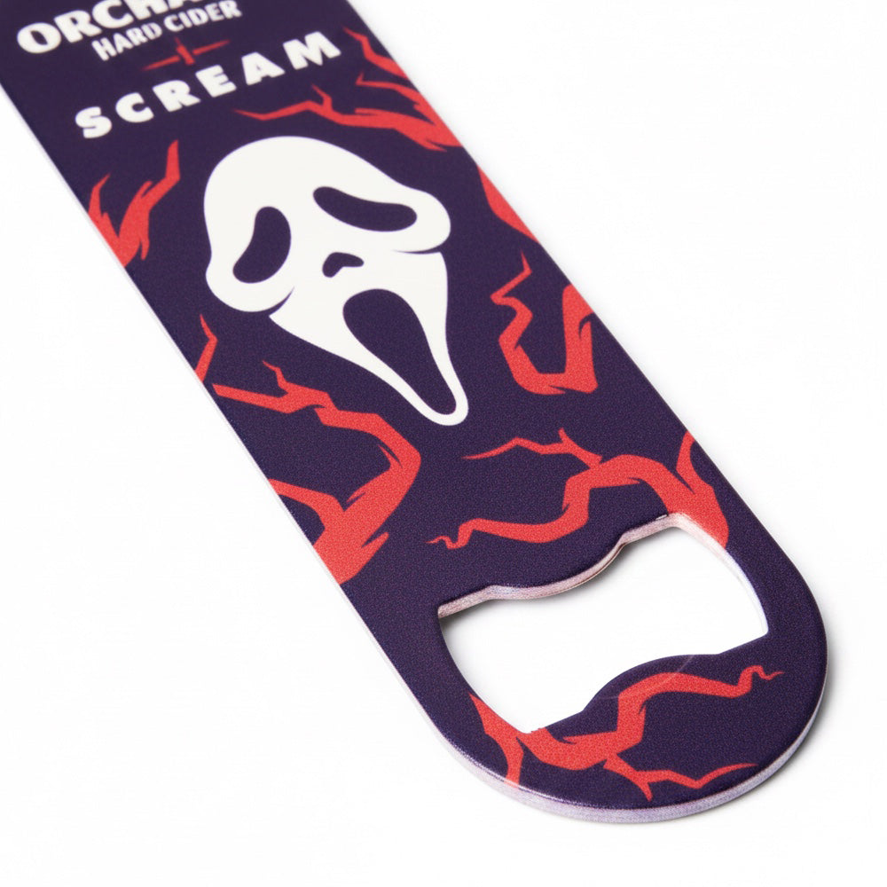 A novelty bottle opener featuring the Ghostface mask from Scream, with 'ANGRY ORCHARD HARD CIDER' and 'SCREAM' printed on a dark purple background with red lightning patterns, shown on a white background.