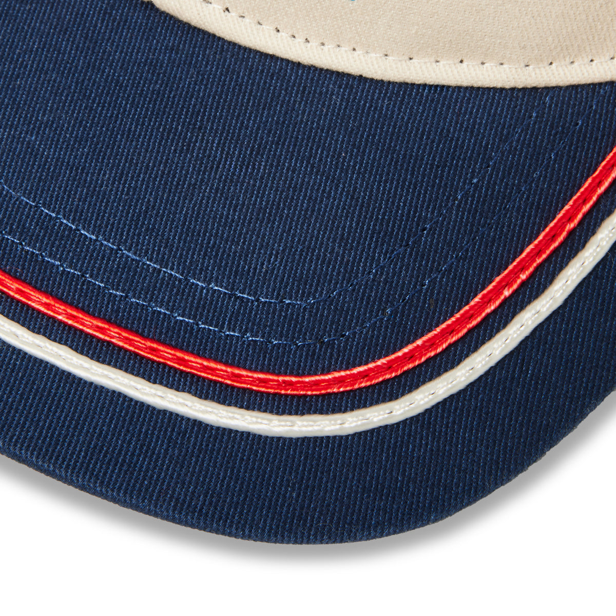 Close-up detail of a baseball cap showing navy blue fabric, a cream-colored top panel, and contrast stitching in red, white, and blue.