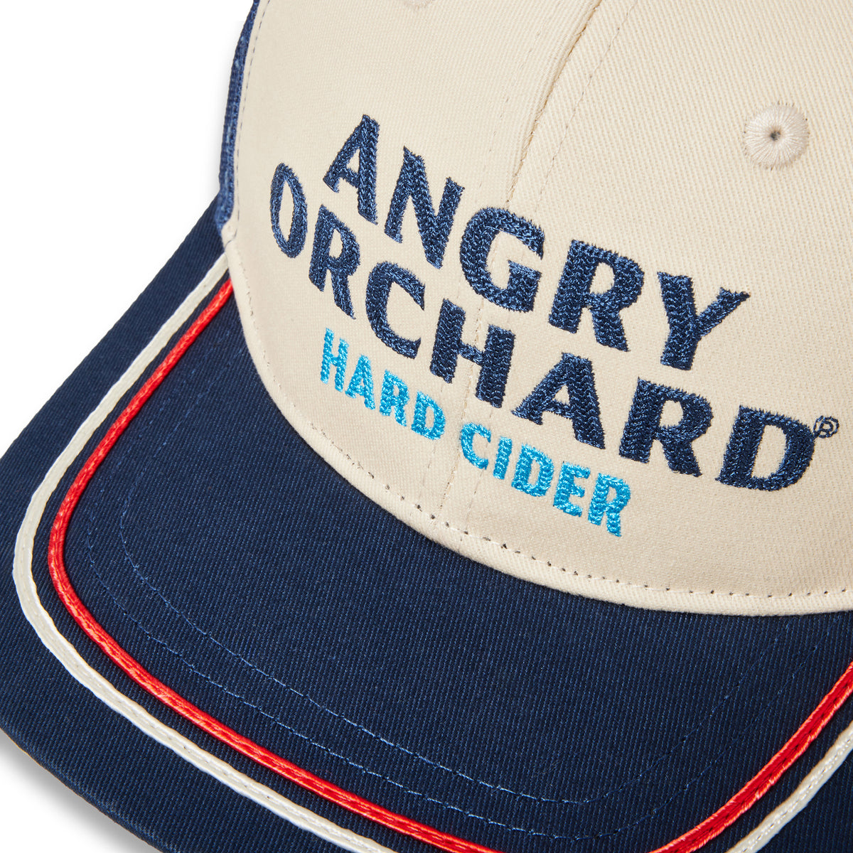 Close-up of a baseball cap with a cream crown, navy blue brim, and embroidered text reading 'ANGRY ORCHARD HARD CIDER' in navy and light blue, with red and white piping along the brim edge.