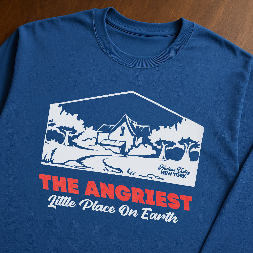 A blue long-sleeve t-shirt laid flat on a dark wood background, featuring a white graphic of a house and trees with the text 'THE ANGRIEST Little Place On Earth' in red and white.