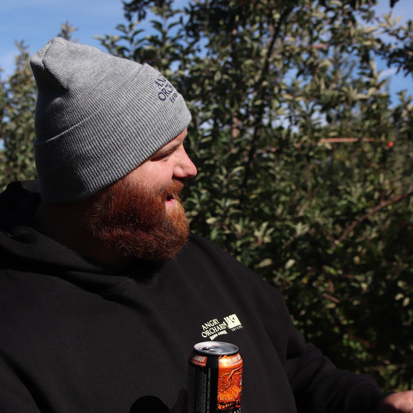 Man holding a blood orange angry orchard can outside, smiling wearing a grey Angry Orchard Hard Cider Carhartt Beanie