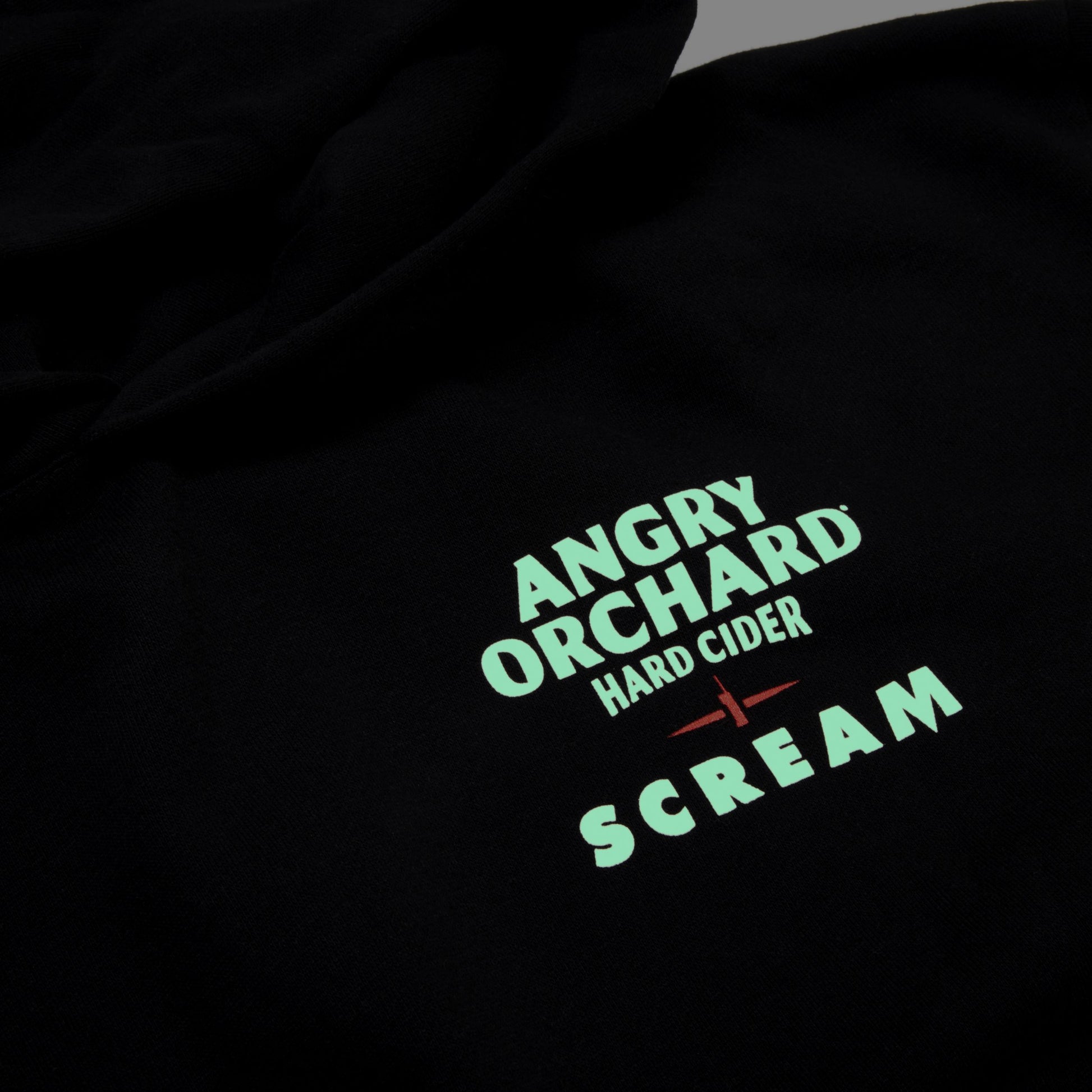Close-up of a black fabric surface with printed text reading 'ANGRY ORCHARD HARD CIDER' and 'SCREAM' in a mint green color, plus a small red accent.