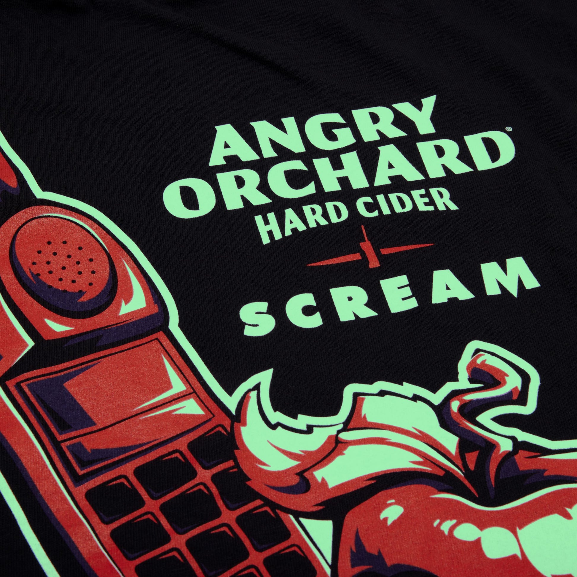 Close-up of a black t-shirt featuring a graphic print of a red rotary phone, a red apple, and neon green text that reads 'ANGRY ORCHARD HARD CIDER SCREAM'.